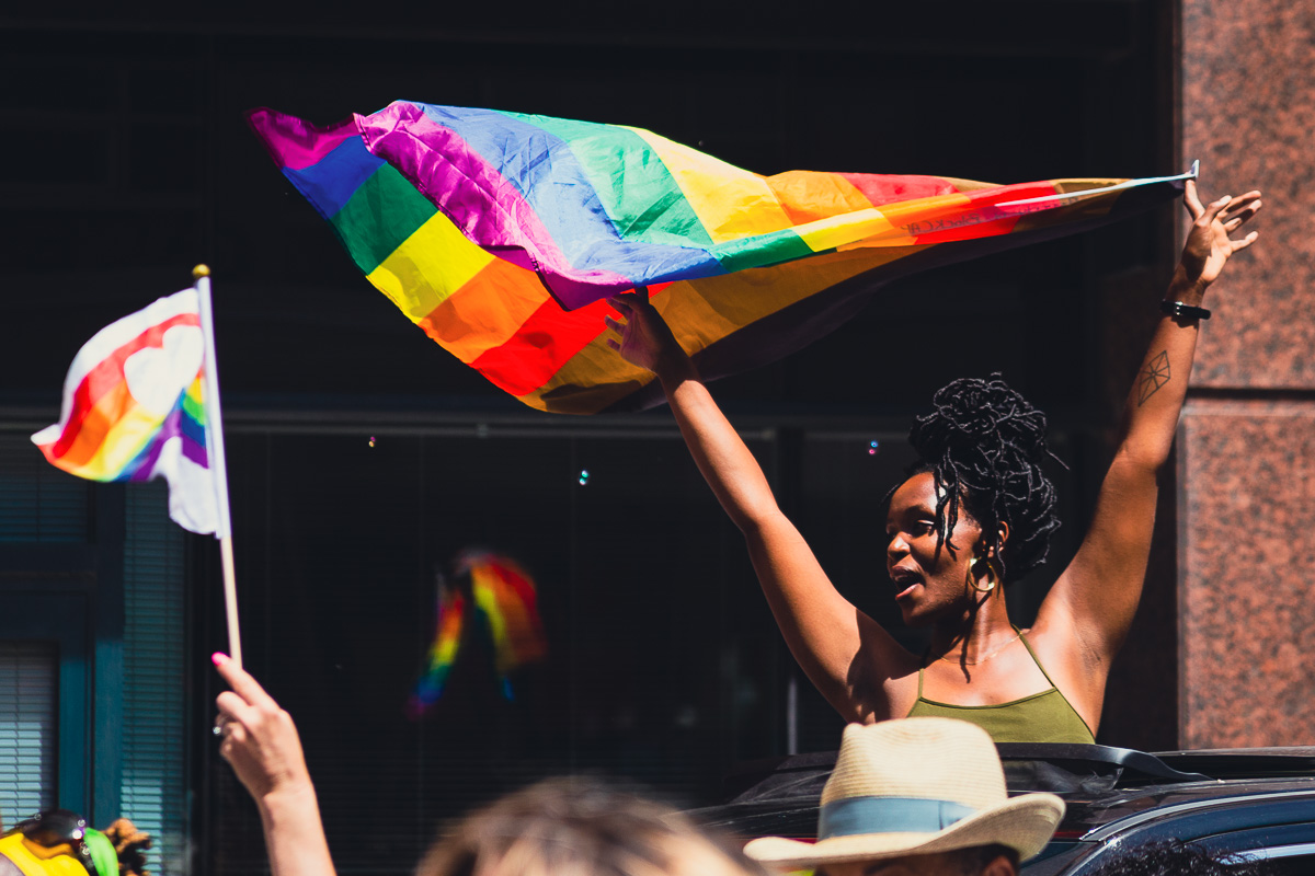 A Black woman standing in a moving car holds a rainbow flag above her head during the Toronto Pride festival’s Dyke March. © Andy Lee, 2022. realandylee.com