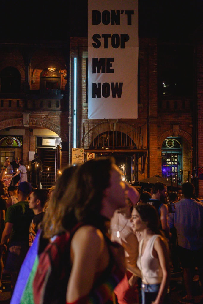 A crowd outside a "Don't Stop Me Now" banner on the street at night. © Andy Lee, 2022. realandylee.com