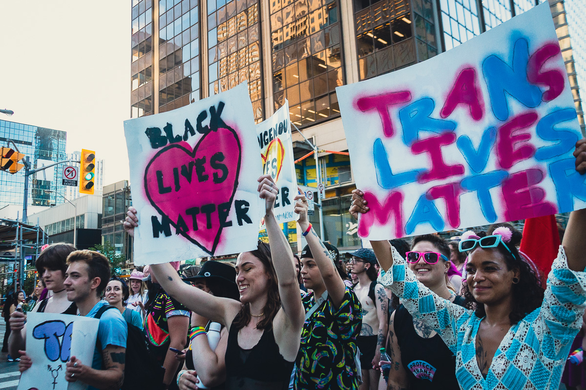 Black woman holding “Trans Lives Matter” sign marching beside white woman holding “Black Lives Matter” sign during Toronto Pride street rally. © Andy Lee, 2022. realandylee.com